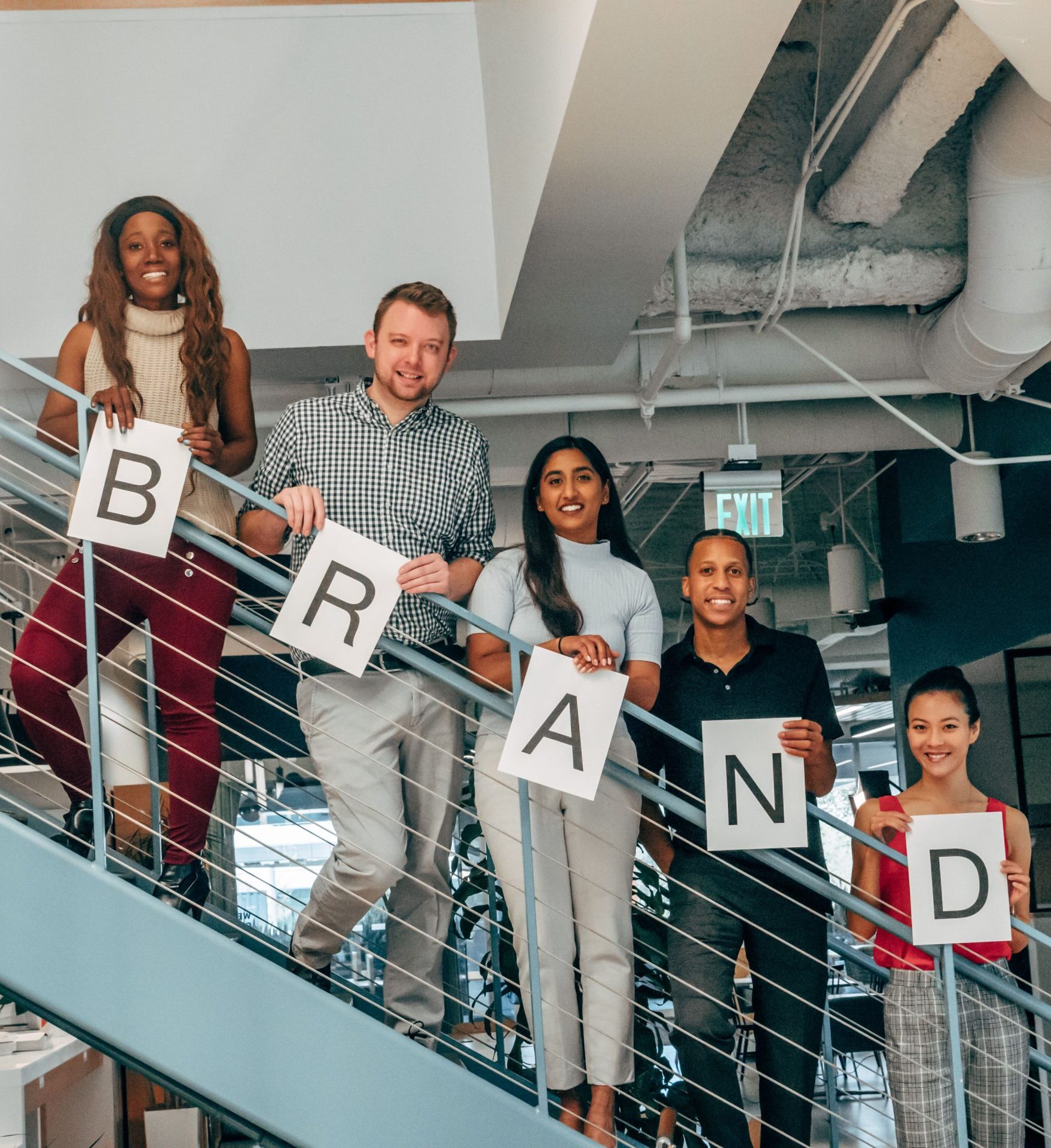 Diverse group of adults smiling and holding 'BRAND' signs on an office staircase.